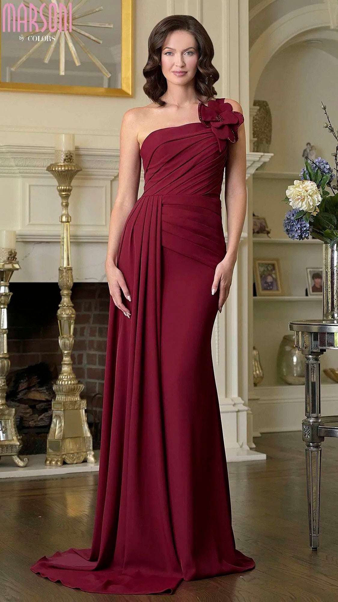 A woman wearing a wine-colored Marsoni M330 long formal dress with a one-shoulder design and 3D floral accent stands indoors by a fireplace and decorative shelves.